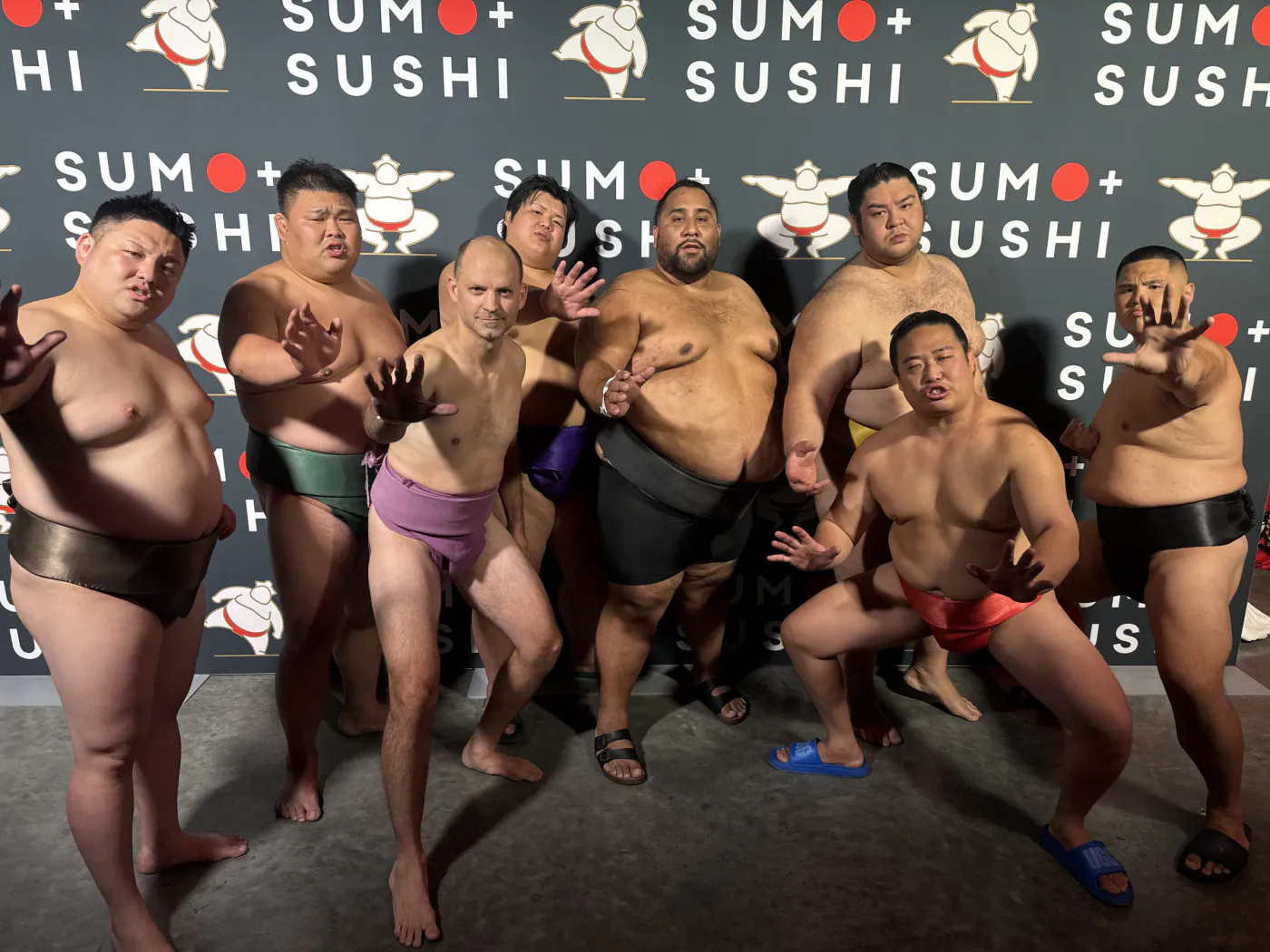 Wrestlers posing with open hands in front of a Sumo and Sushi backdrop
